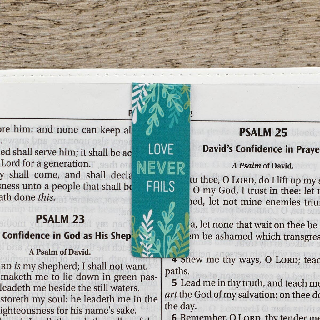 Keepsake lane teal bookmark with white leaf pattern and the inspirational text Love Never Fails resting on a Bible